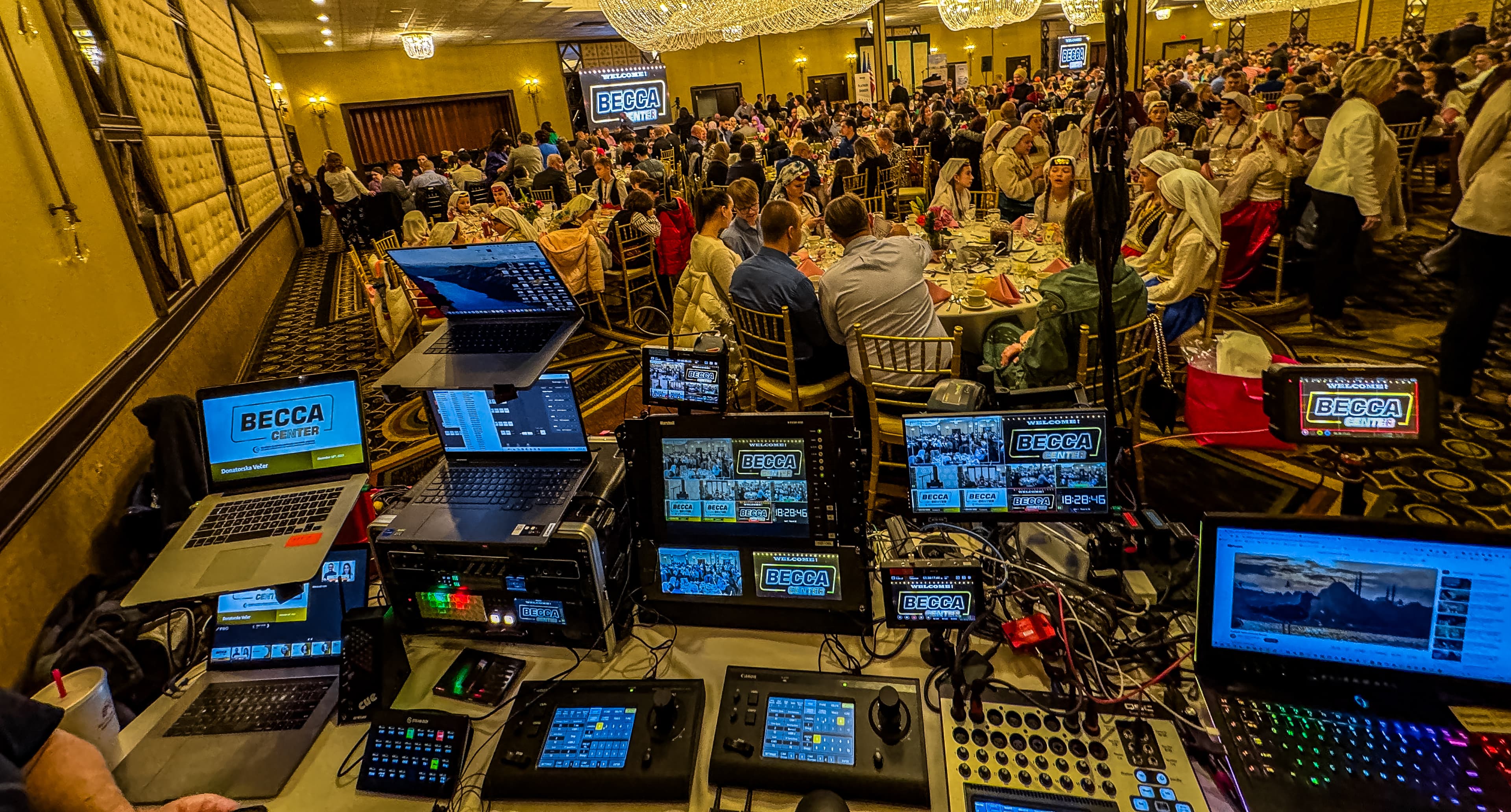 Video production equipment monitors a crowded banquet hall event with Becca Center branding on screens.