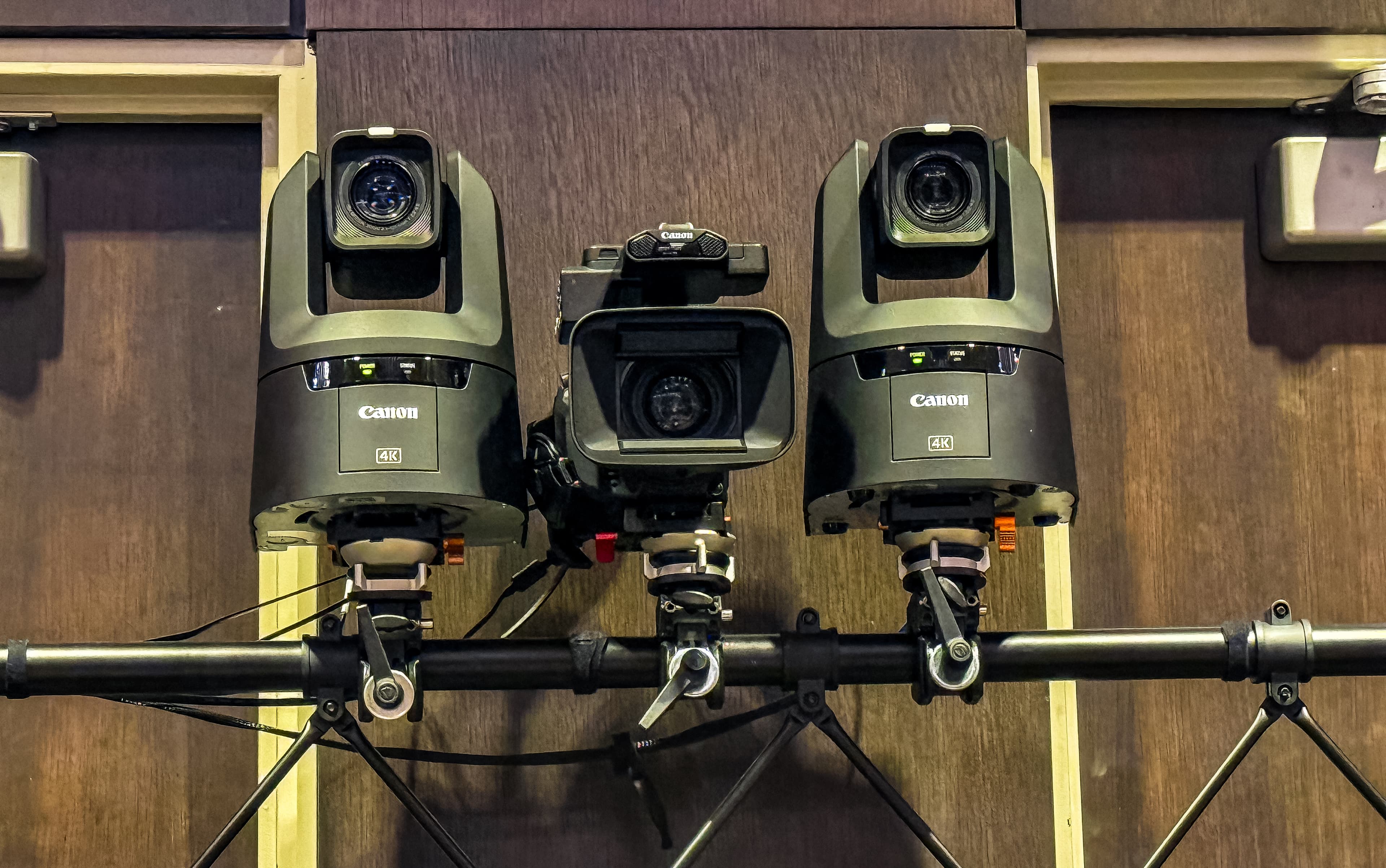 Three professional Canon 4K cameras mounted on a horizontal bar against dark wood paneling.