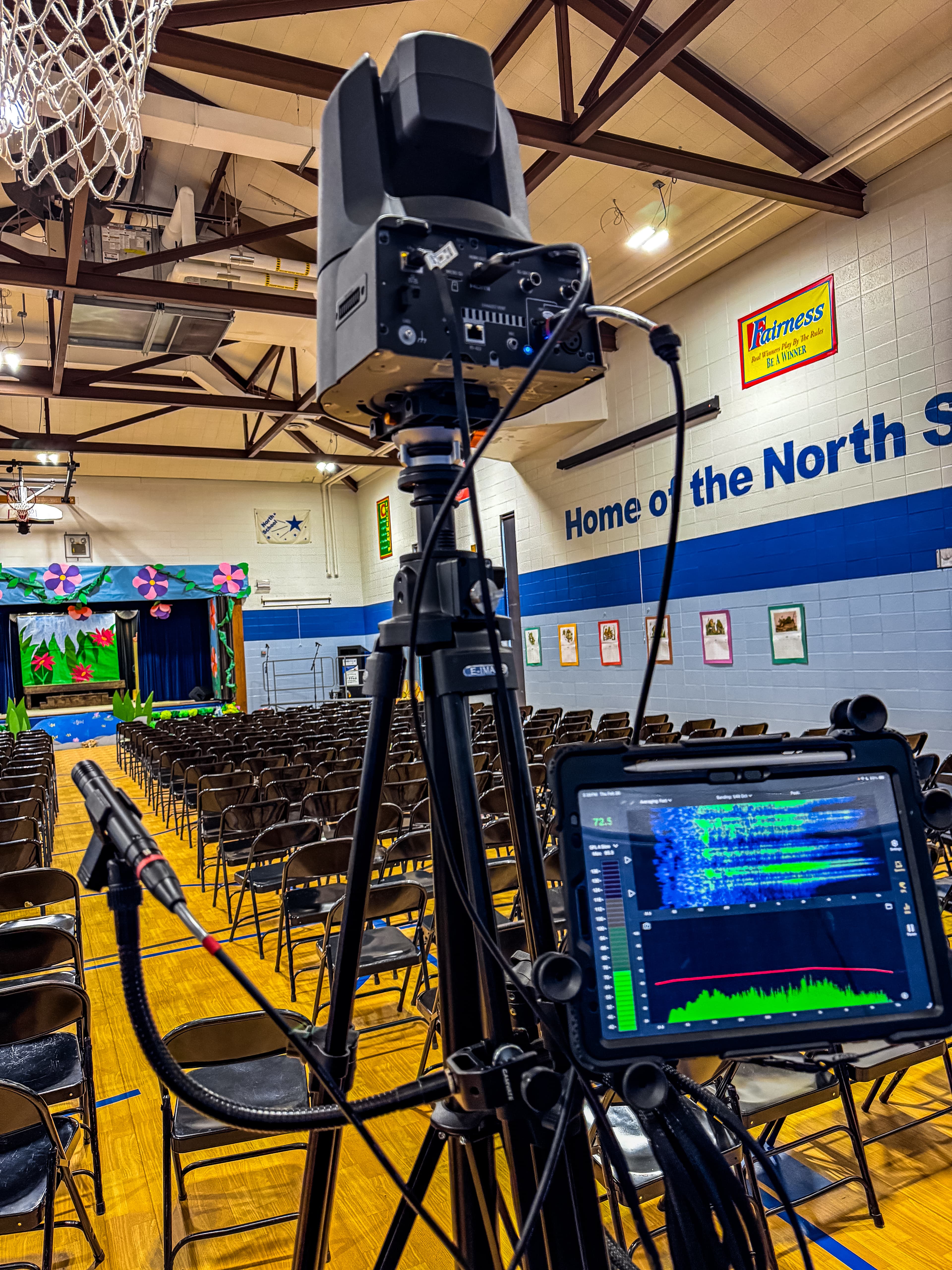 Professional camera and tablet on a tripod in an empty school gym facing a stage.