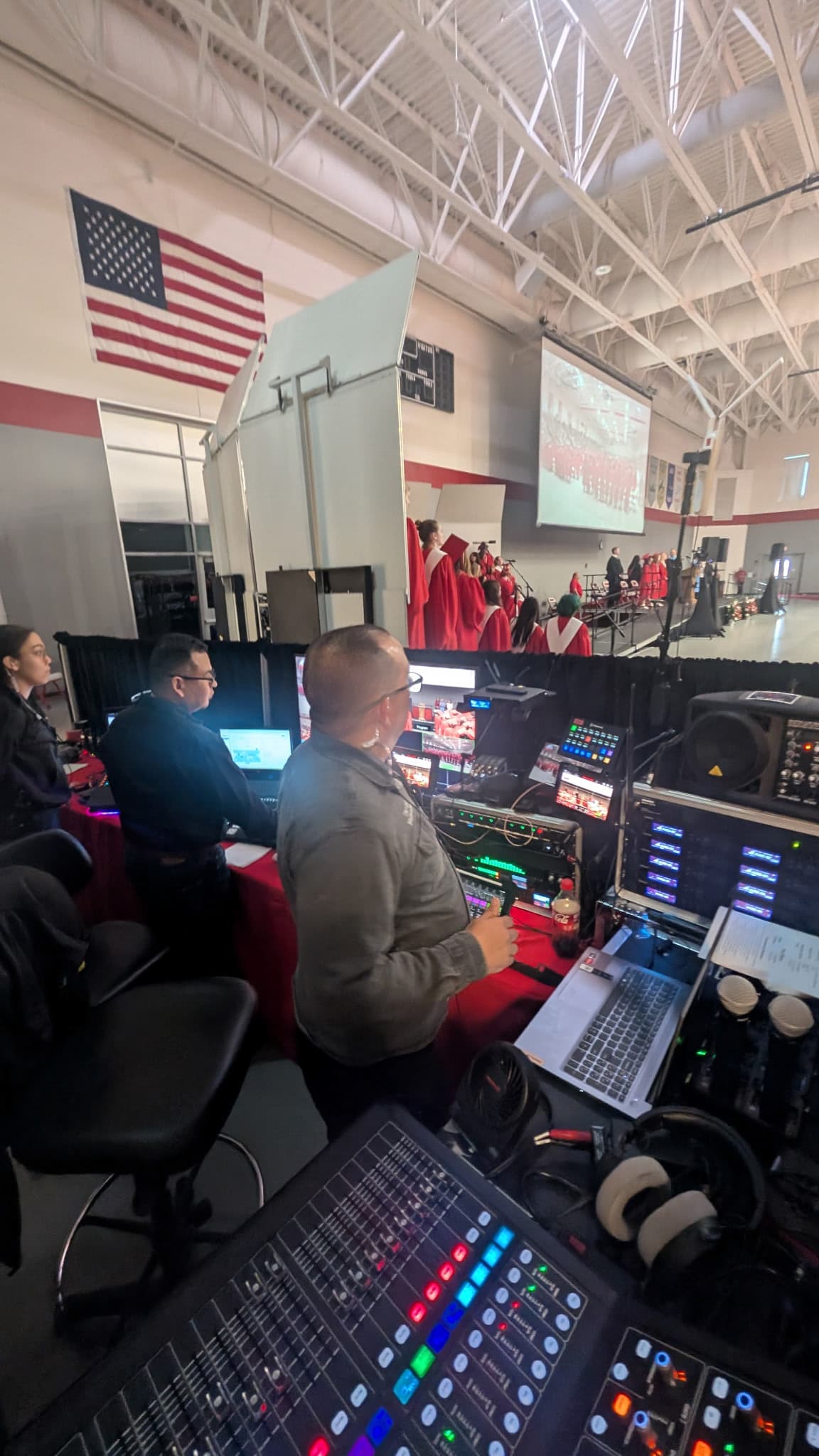 AV technicians operate soundboards and monitors during a graduation ceremony in a school gymnasium.